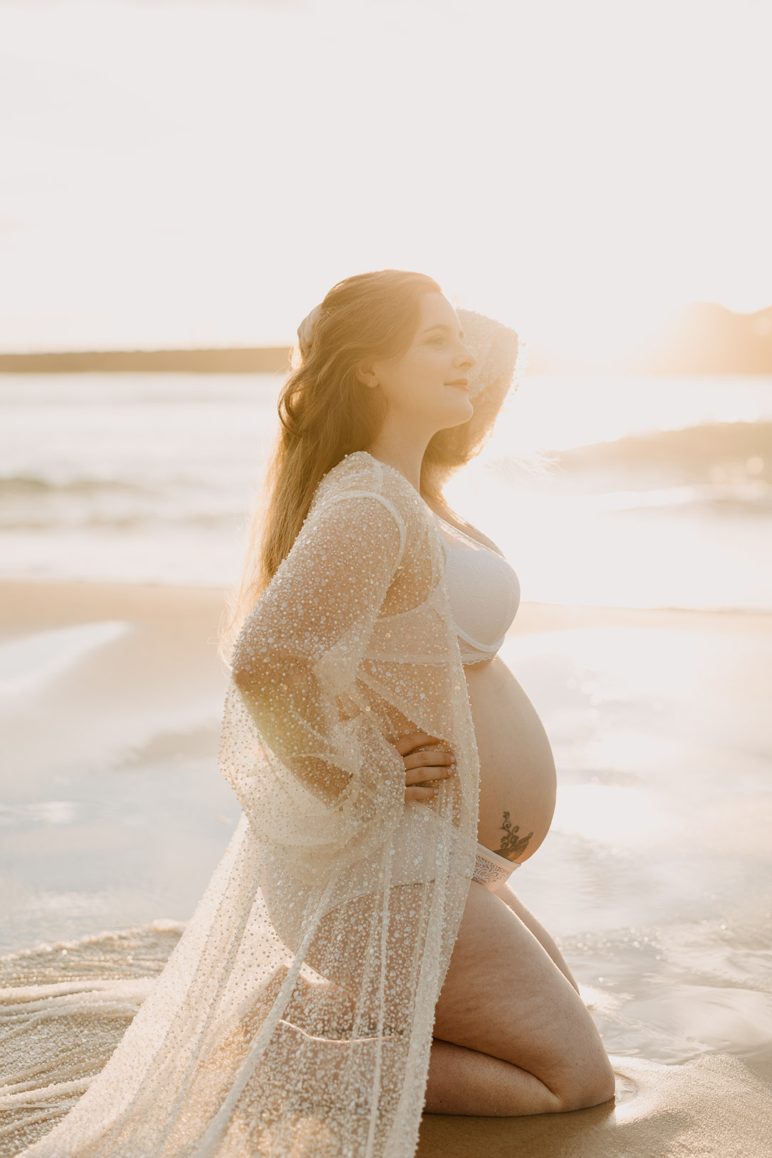 A pregnant mum wearing a sheer white robe kneels on a wet sandy beach, the golden rays of sunrise illuminate her bump. The photo was taken by Studio Magnolia, a Brisbane maternity photographer.